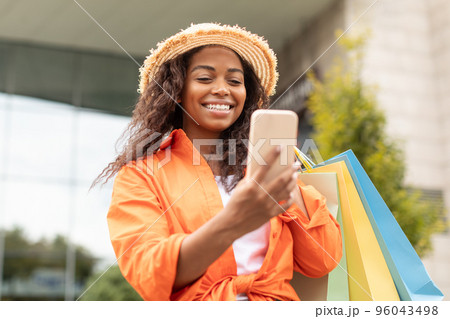 Smiling black lady with many packages with purchases has video call on phone, enjoys talk in city 96043498