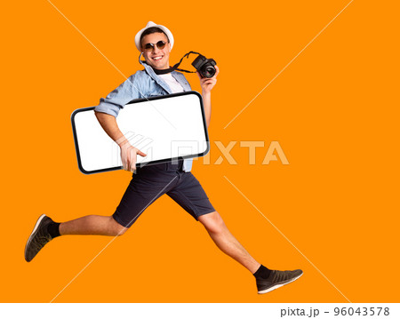 Cheerful Male Tourist Holding Camera Jumping With Big Blank Smartphone In Hand Cheerful Male Tourist Holding Camera Jumping With Big Blank Smartphone In Hand 96043578