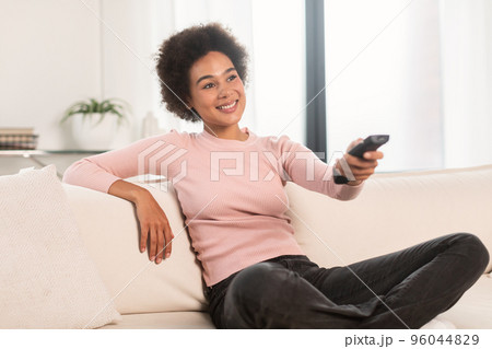 Smiling relaxed millennial african american woman with remote control watching tv sit on sofa, enjoy free time 96044829