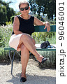 A beautiful middle-aged woman in her 50s, in a short skirt, bare legs, in a summer t-shirt, with sunglasses, on a nice sunny day, sitting on a park bench 96046001