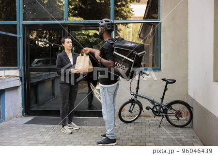Office food delivery service, worker taking order package. African american courier delivering restaurant takeaway meal, giving employee paper bag near company building outdoors 96046439