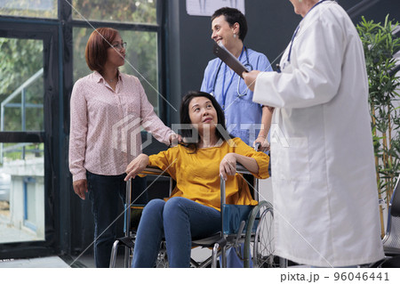 Senior doctor talking to wheelchair user and consulting injured patient sitting in waiting area. Physician giving support and advice to patient with illness, checkup visit. Medicine support 96046441