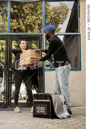 Pizzeria courier passing woman pizza boxes big pile, man taking out order from thermal bag in front of office building. Food delivery service, african american deliveryman giving customer meal 96046496