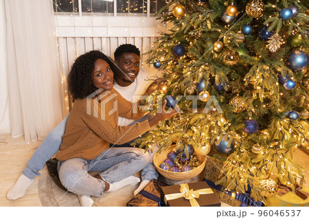 Glad young african american lady and guy decorate Christmas tree with garlands, toys in cozy living room 96046537
