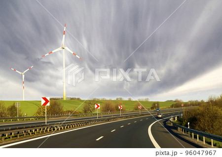 Wide highway with moving car stretching to horizon by green hills with high wind turbines on blue cloudy sky background. Modern technologies, renewable energy source and ecological problems solving. Wide highway with moving car stretching to horizon by green hills with high wind turbines on blue cloudy sky background. Modern technologies, renewable energy source and ecological problems solving. 96047967