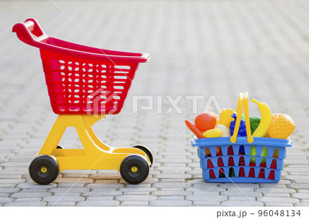 Shopping pushcart and a basket with toy fruits and vegetables. Bright plastic colorful toys for children outdoors on sunny summer day. Shopping pushcart and a basket with toy fruits and vegetables. Bright plastic colorful toys for children outdoors on sunny summer day. 96048134
