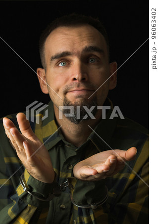 Bearded man handcuffed with palms apart. Caucasian man in green plaid shirt with cuffs on a black background looking at the camera. Facial expression is played out. Bearded man handcuffed with palms apart. Caucasian man in green plaid shirt with cuffs on a black background looking at the camera. Facial expression is played out. 96063402
