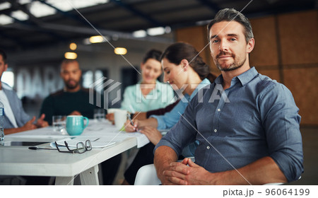 The creative process. Portrait of a mature man sitting at a table in an office with colleagues working in the background. 96064199