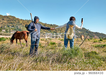 Back view of couple holding hands while walking on rural meadow with horses grazing on autumn or spring pasture. Woman takes a photo with smartphone. Unity with nature. Road to mountain. 96066430