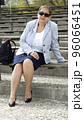A middle-aged busty woman in grey jacket, in pantyhose, nylons, with sunglasess sitting on a bench, outdoor, on a sunny day 96066451