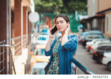 An elegant woman in summer clothes is talking on the phone straightening her hair. Street with cars in the background. Concept of communication and social networks 96068785