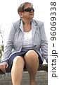 A middle-aged busty woman in grey jacket, in pantyhose, nylons, with sunglasess sitting on a bench, outdoor, on a sunny day 96069338