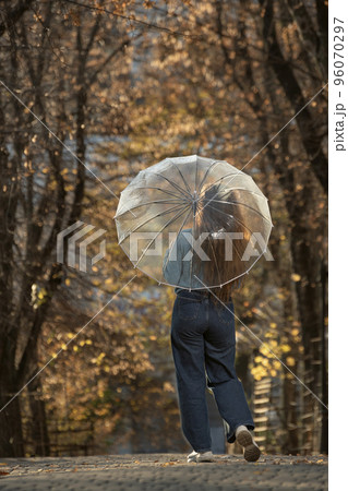 Brunette with long hair in blue sweater and pants stands with back under transparent umbrella in autumn park Brunette with long hair in blue sweater and pants stands with back under transparent umbrella in autumn park 96070297