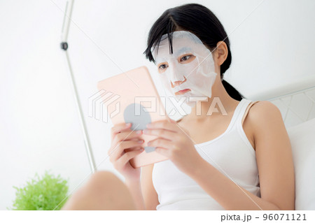 Beautiful young asian woman with sheet facial mask sitting watching tablet computer on bed. 96071121