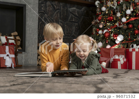 Two blond siblings lie on floor under Christmas tree. Smiling brother and sister reads the book. Christmas eve 96071532