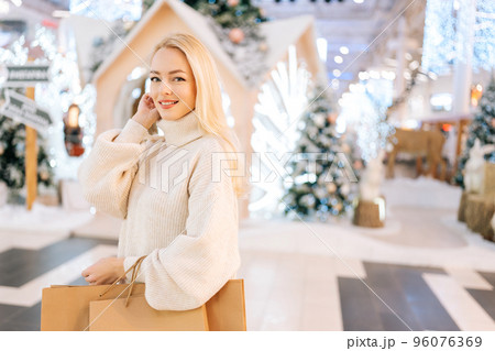Portrait of charming young woman with blonde hair holding bags with purchase smiling looking at camera standing in hall of celebrate shopping mall in Christmas eve, on background of xmas decorations. 96076369