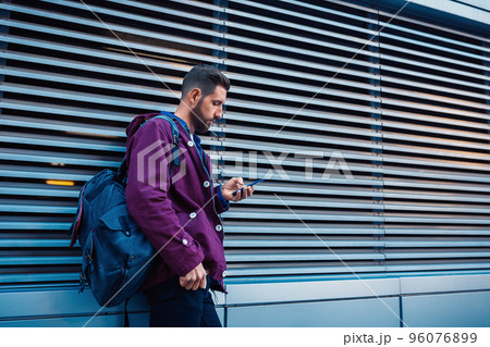 Confident bearded man holding in and mobile phone while standing near street wall in sunny summer evening. 96076899