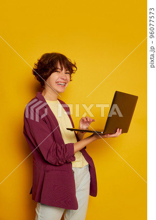 Portrait of cheerful teen girl, student with curly brown short hair posing with laptop isolated over yellow background 96077573