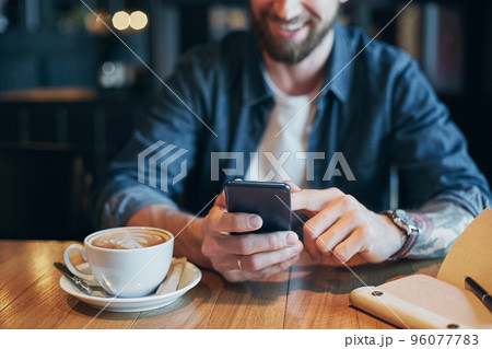 Man hands in denim shirt slide with finger on screen his smart phone, near cup with coffee on wooden table Man hands in denim shirt slide with finger on screen his smart phone, near cup with coffee on wooden table 96077783