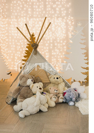 Children's photo zone in the Christmas photo studio - wigwam with teddy bears on the background of silhouettes of fir trees and garlands. Children's photo zone in the Christmas photo studio - wigwam with teddy bears on the background of silhouettes of fir trees and garlands. 96080710