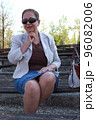 A middle-aged woman in a blue skirt and gray jacket, slightly crossed legs, in pantyhose, and with sunglasses, sits on a bench and looks at the camera as if she wants to say something 96082006