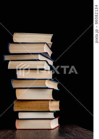 Tall stack of hardback books on a wooden surface against a dark background. Tall stack of hardback books on a wooden surface against a dark background. 96082134