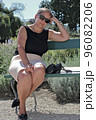 A middle-aged sexy woman in a summer skirt and T-shirt slightly crossed bare legs, and sunglasses sits on a bench in the park with a pensive look and looks at the camera 96082206