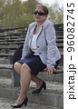A middle-aged busty woman in grey jacket, in pantyhose, nylons, with sunglasess sitting on a bench, outdoor, on a sunny day and looks at the camera 96082745