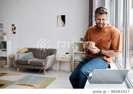 Happy young man with cup of coffee sitting in front of laptop by window 96083927