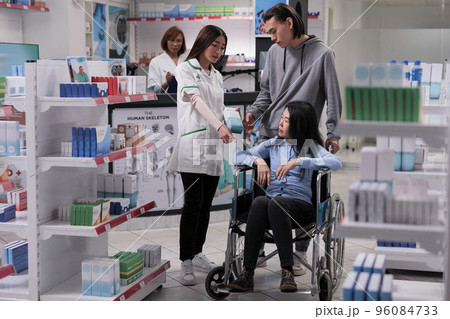 Young man and adult in wheelchair buying medicine, receiving medical help and advice from pharmacist in drugstore. Customer dealing with physical impairment in pharmacy, buy pills. Young man and adult in wheelchair buying medicine, receiving medical help and advice from pharmacist in drugstore. Customer dealing with physical impairment in pharmacy, buy pills. 96084733