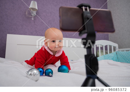 A baby boy dressed in a Santa suit sits on the bed and looks at the sick phone. Christmas greetings by video call 96085372