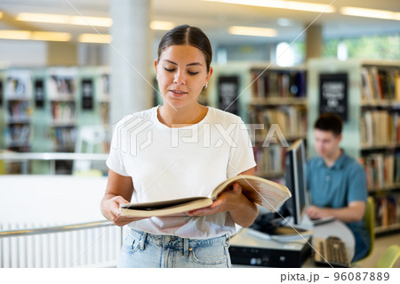 Smiling student girl standing in the university library, holding textbooks in hands 96087889
