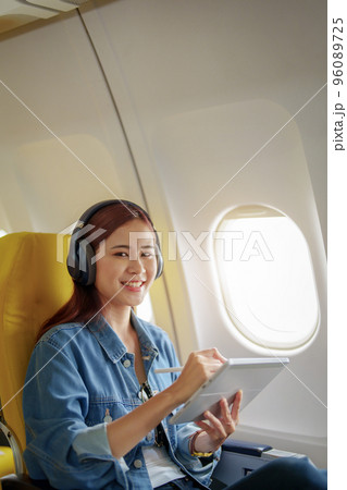 Attractive portrait of an Asian woman sitting in a window seat in economy class using tablet and listening to instrumental music during an airplane flight, travel concept, vacation, relaxation. 96089725