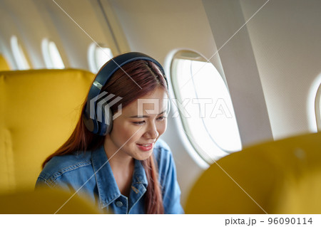 Attractive portrait of an Asian woman sitting at a window seat in economy class listening to music during a flight on a plane, travel concept, vacation, relaxation. 96090114