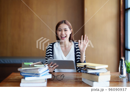 A portrait of a young Asian woman with a smiling face using a tablet computer during an online video conferencing class in a library. A portrait of a young Asian woman with a smiling face using a tablet computer during an online video conferencing class in a library. 96090937