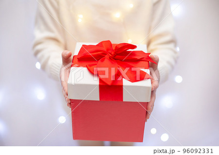 Child in white sweater holding red Christmas gift in hand on light bokeh background 96092331