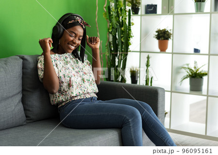 African woman listening to music with headphones while sitting on couch at home 96095161