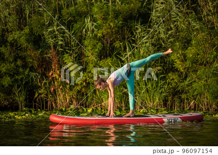 Photo of young woman doing hand stand on stand up paddle board. She wearing a leggings and top. 96097154