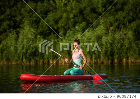 Young athletic woman doing fitness on a board with an oar on a lake. 96097174