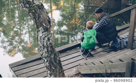 High angle view of people father and son fishing in pond in forest sitting on chairs on wooden pier and holding rods. Family, common hobby and nature concept. High angle view of people father and son fishing in pond in forest sitting on chairs on wooden pier and holding rods. Family, common hobby and nature concept. 96098902
