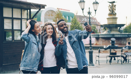 Happy African American guy is taking selfie with friends beautiful Caucasian girls standing in street posing and holding modern smartphone during journey in foreign country. Happy African American guy is taking selfie with friends beautiful Caucasian girls standing in street posing and holding modern smartphone during journey in foreign country. 96099017