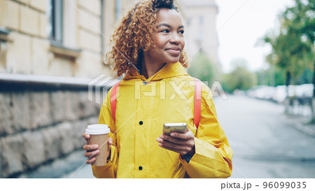 Smiling African-American girl is using smartphone texting friends and holding to-go coffee walking in city alone. Modern technology, communication and take away drinks concept. 96099035