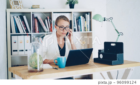 Confident young woman company employee is talking on mobile phone, using laptop and taking notes in notebook sitting at desk at workplace. Business, modern office and people concept. Confident young woman company employee is talking on mobile phone, using laptop and taking notes in notebook sitting at desk at workplace. Business, modern office and people concept. 96099124