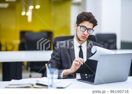 Young office worker in glasses reads documents in the office. Stylish man analyze documents, plans. Young office worker in glasses reads documents in the office. Stylish man analyze documents, plans. 96100716