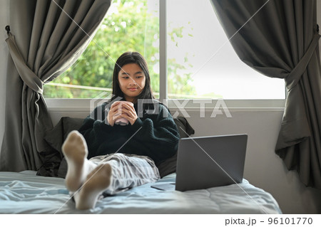 Smiling asian girl in warn clothes drinking hot chocolate and using laptop in bedroom 96101770