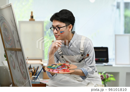 Pleased male artist holding palette and looking at his artwork with admiration 96101810