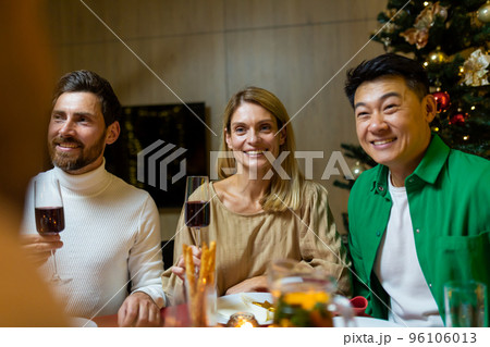Happy young interracial people sitting at festive New Year's table. They are holding glasses of wine, talking, smiling, celebrating Christmas with friends. Happy young interracial people sitting at festive New Year's table. They are holding glasses of wine, talking, smiling, celebrating Christmas with friends. 96106013
