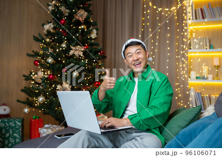 Portrait of a young handsome Asian man sitting on the sofa at home near the Christmas tree in a red hat, holding a laptop and a phone, looking at the camera, smiling, pointing super. 96106071
