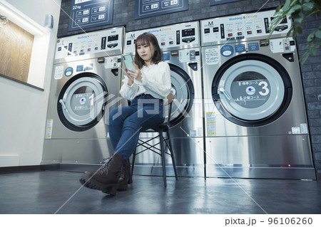 コインランドリーで洗濯が終わるのを座ってスマホを見ながら待つ若い女性 96106260
