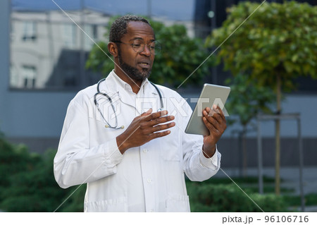 Smiling senior mature doctor in medical coat and glasses with tablet talking on video call remotely online, african american man outside clinic consulting patients. 96106716